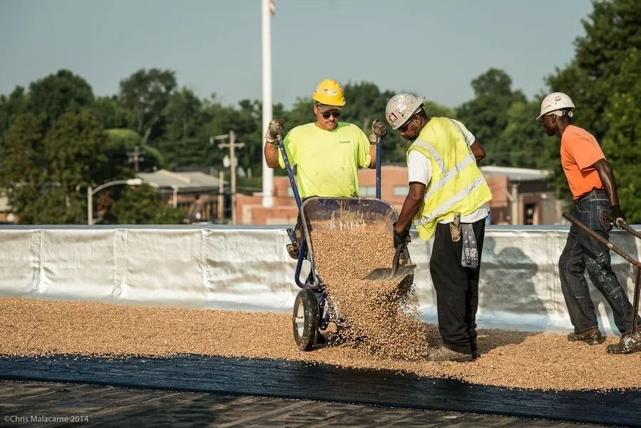 Three construction workers wearing hard hats and safety gear spread gravel across a flat roof surface.