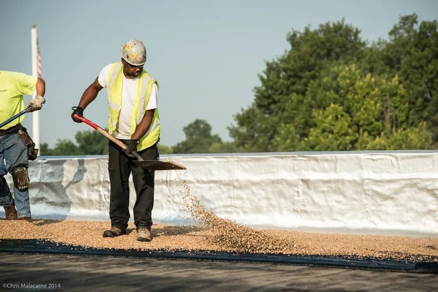 Construction workers spread gravel over a black surface on a rooftop against a clear sky and trees.