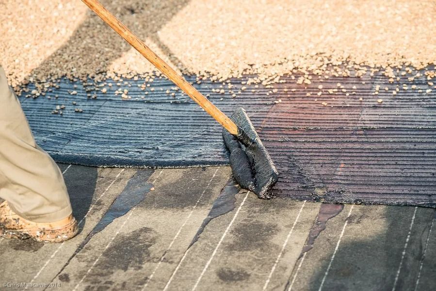 A person uses a long-handled squeegee to spread black adhesive across a substrate while applying gravel over the top.