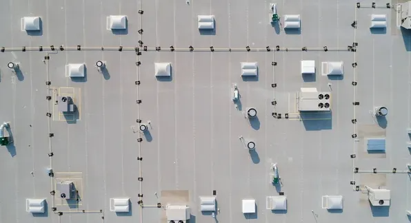 An aerial view of a flat, light-gray industrial roof featuring numerous ventilation units, skylights, and piping.