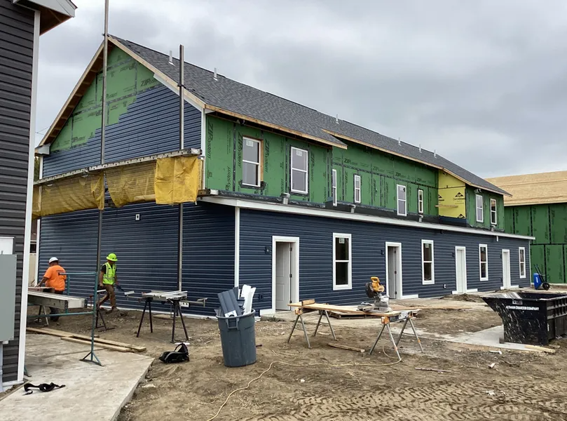 Construction workers stand outside a partially completed navy blue residential building with exposed green sheathing.