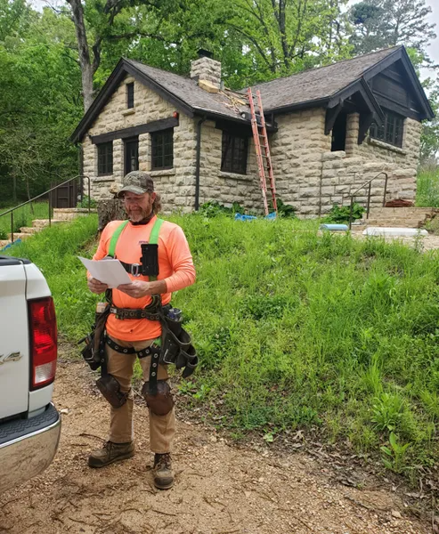 A worker in high-visibility gear reads a document near a stone cottage with a ladder against the roof.
