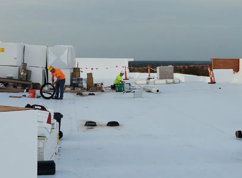 Construction workers in high-visibility gear work on a white flat roof surrounded by materials and safety barriers.