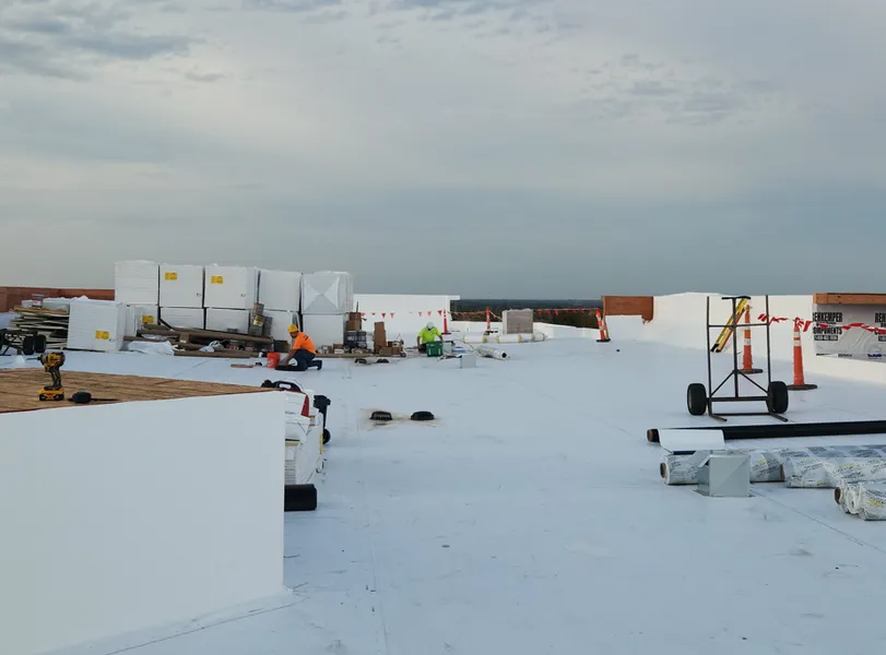 Construction workers on a roof covered in white membrane, with stacks of building materials and equipment nearby.