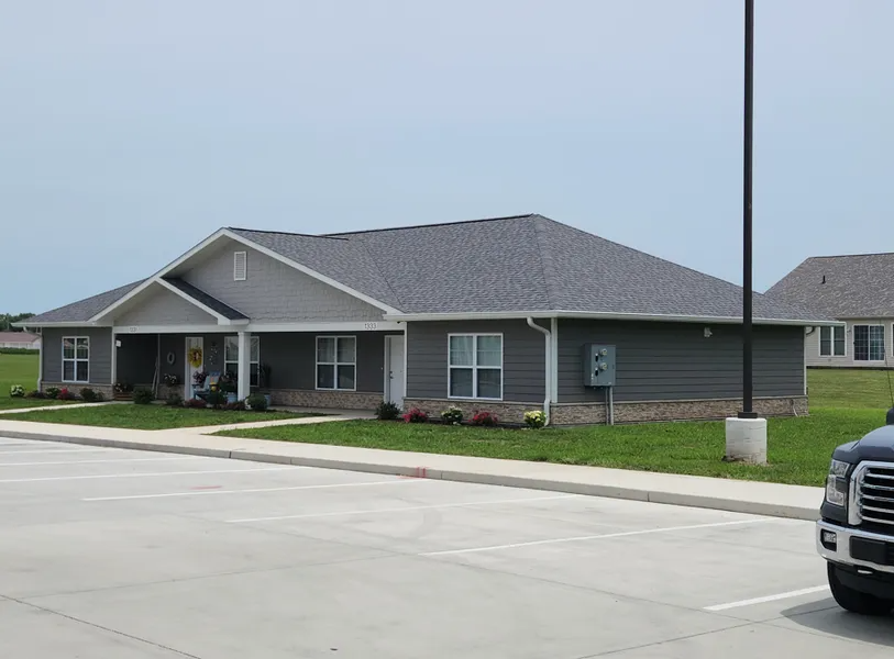 A gray, single-story residential building with a brick base and a parking lot in the foreground under a clear sky.