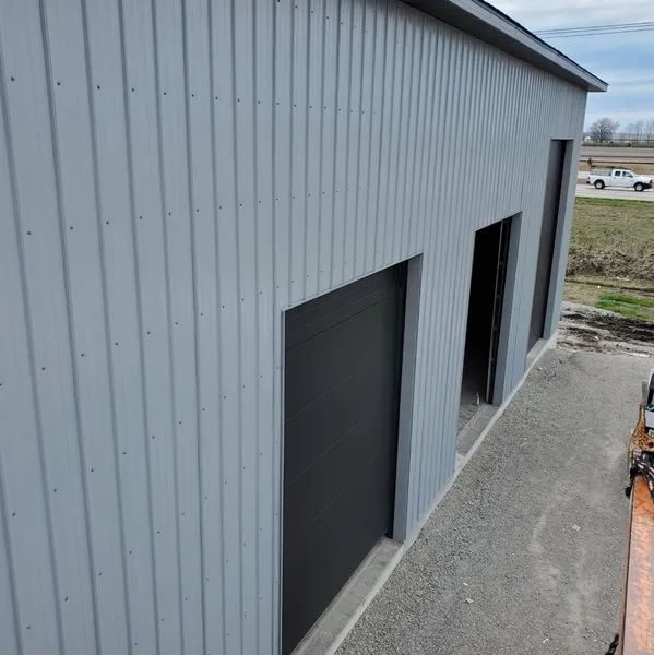 A high-angle view of a grey metal building featuring two black garage-style doors and one open doorway.