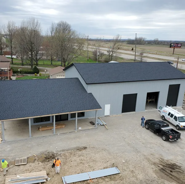 Aerial view of a gray metal building under construction with a dark shingle roof, a front porch, and workers on-site.