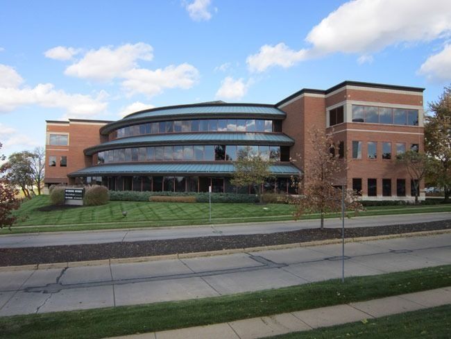 A three-story office building with red brick walls, horizontal windows, and a curved glass facade under a cloudy blue sky.