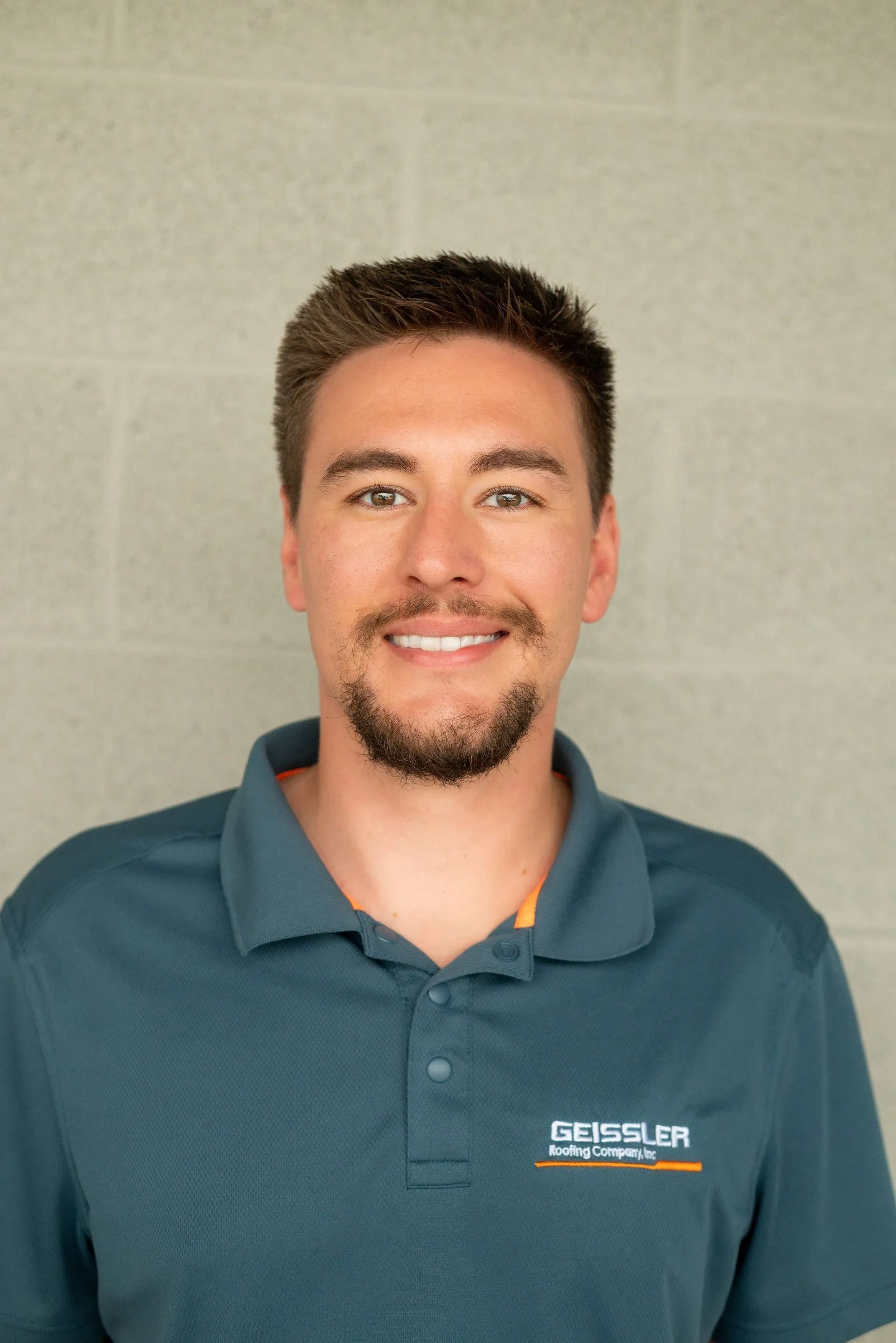 A person with a short beard smiles while wearing a teal polo shirt with a logo, positioned against a gray block wall.