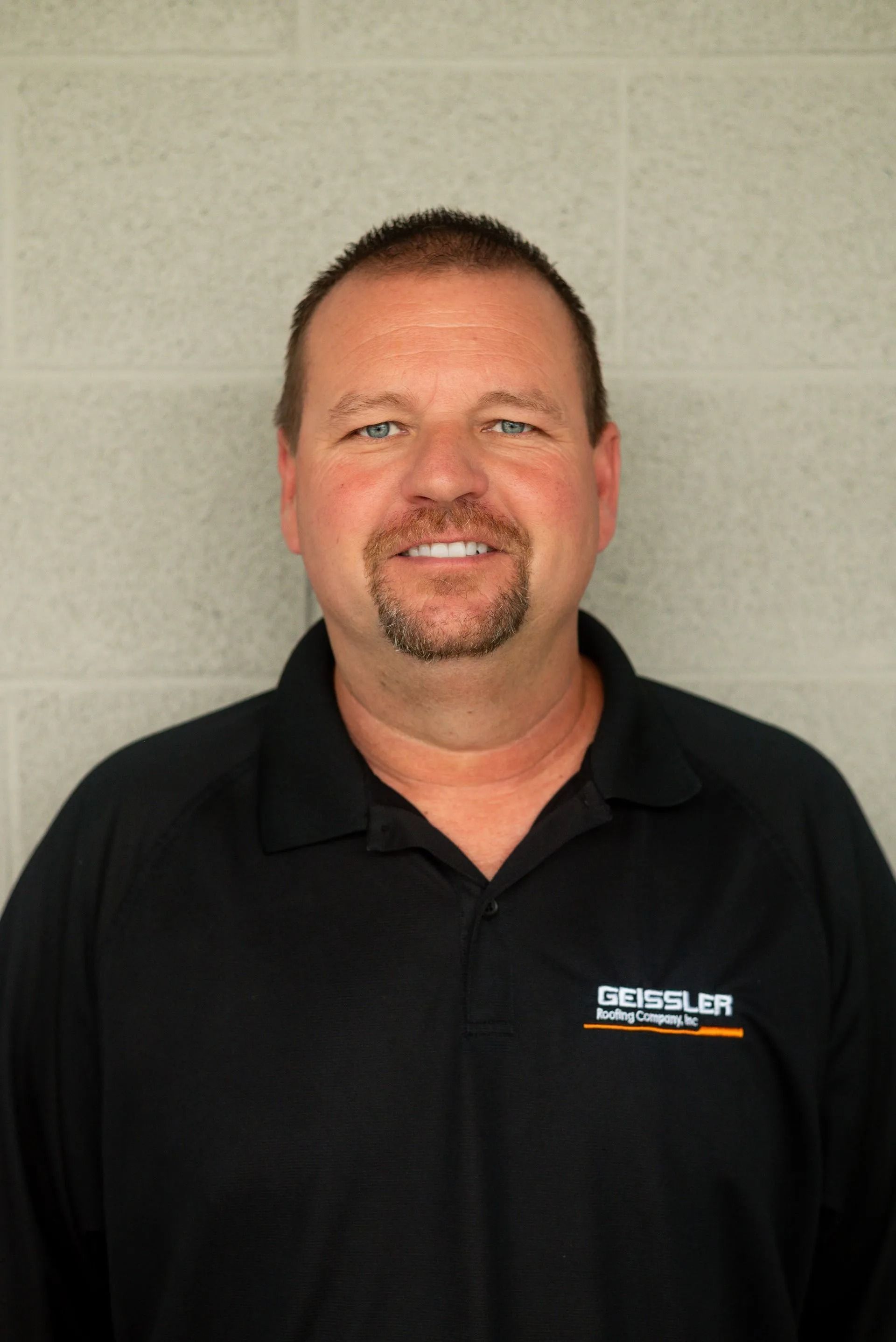 A smiling person with a goatee wearing a black polo shirt with a company logo, standing before a gray block wall.
