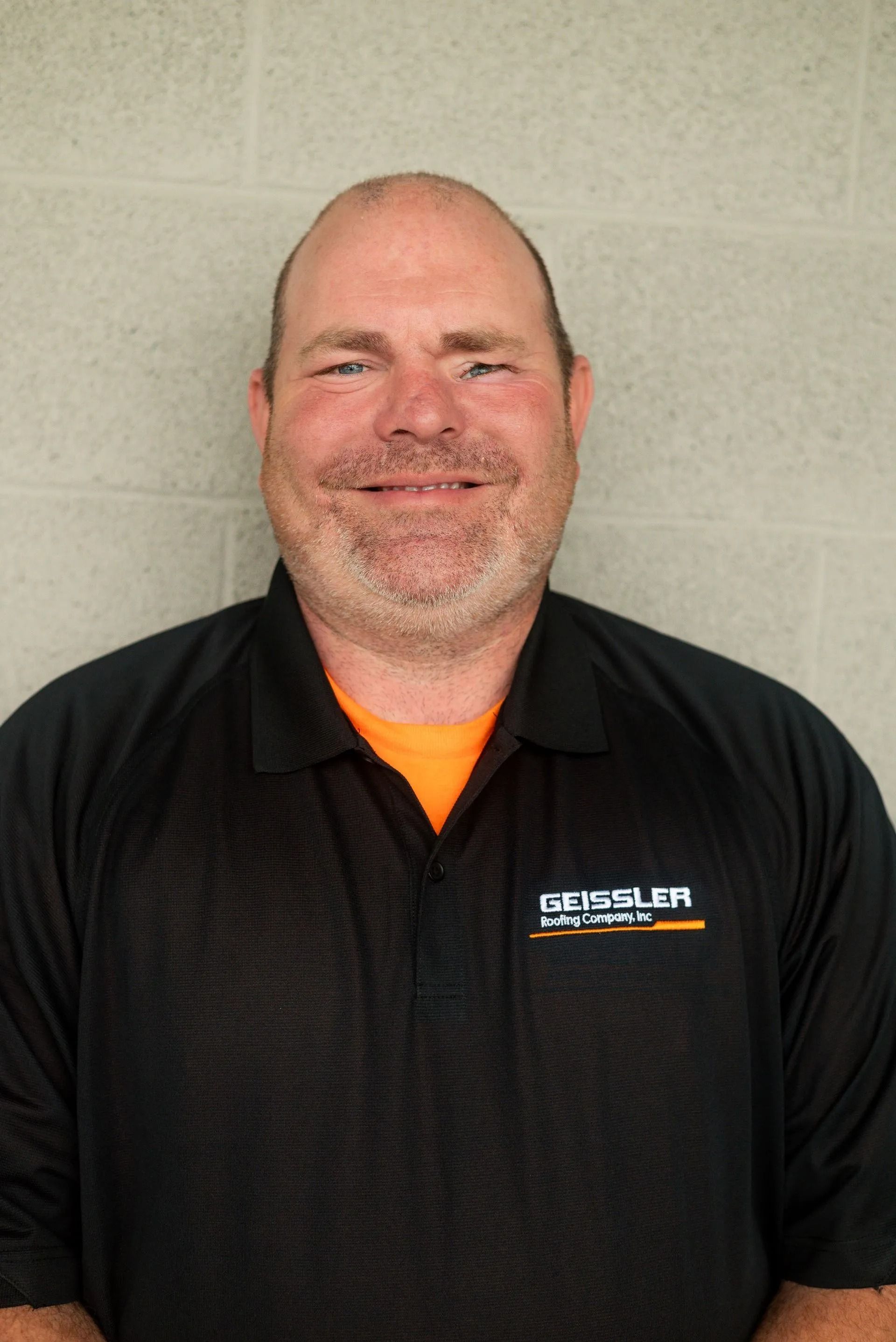 A person with a shaved head and light facial hair, wearing a black collared shirt with an orange under-shirt.