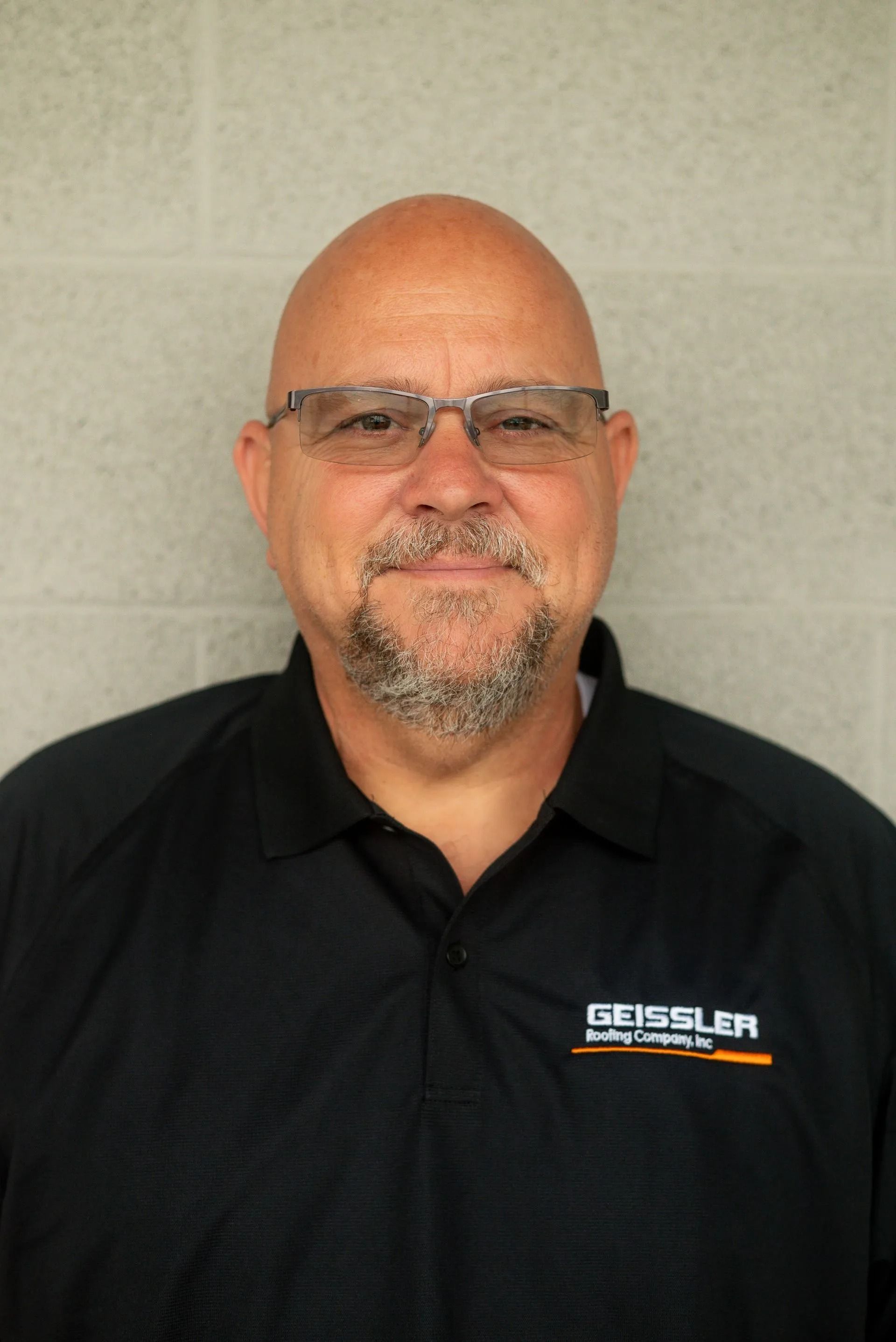 A smiling person wearing glasses and a black branded polo shirt stands in front of a grey cinder block wall.