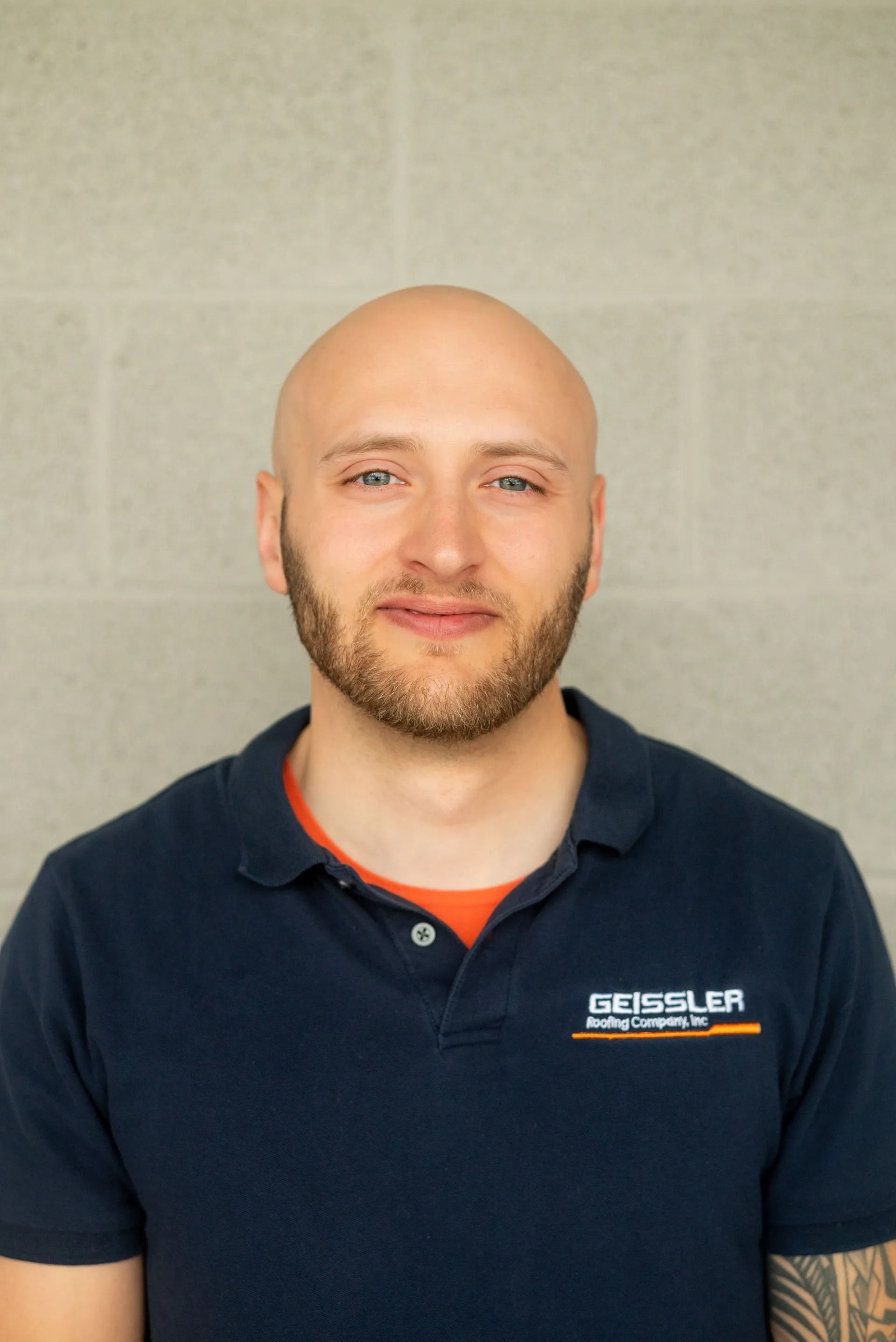 A bald person with a beard, wearing a navy blue polo shirt with an orange undershirt, against a light gray brick wall.