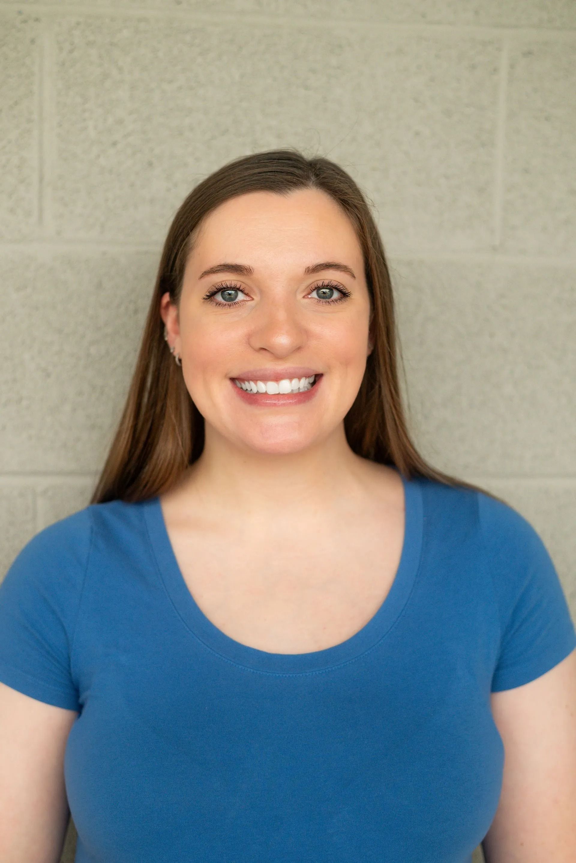 A person with long brown hair, wearing a blue shirt, smiling against a light gray brick wall background.