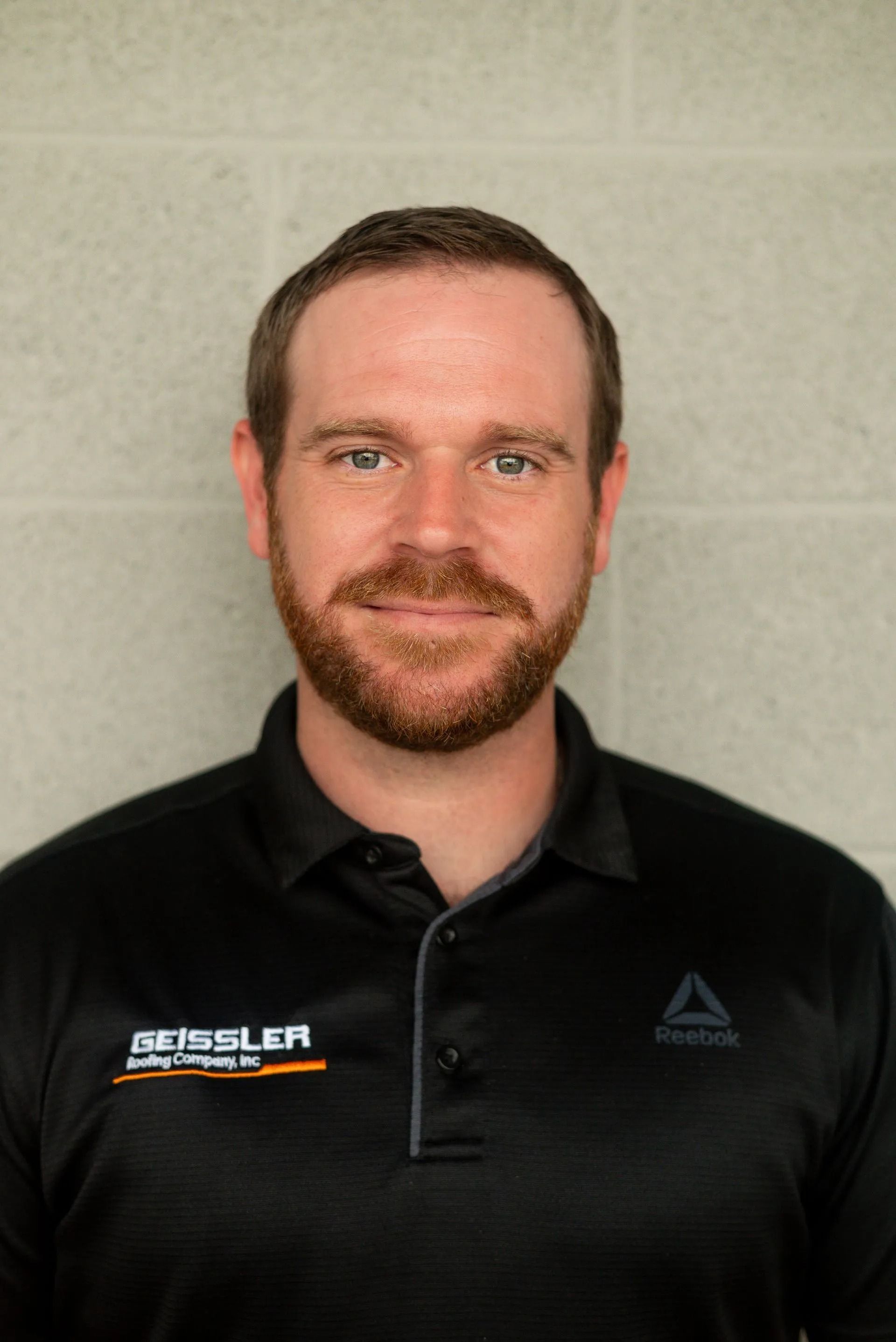 A person with a beard, wearing a black Reebok polo shirt with a company logo, posing against a light gray brick wall.
