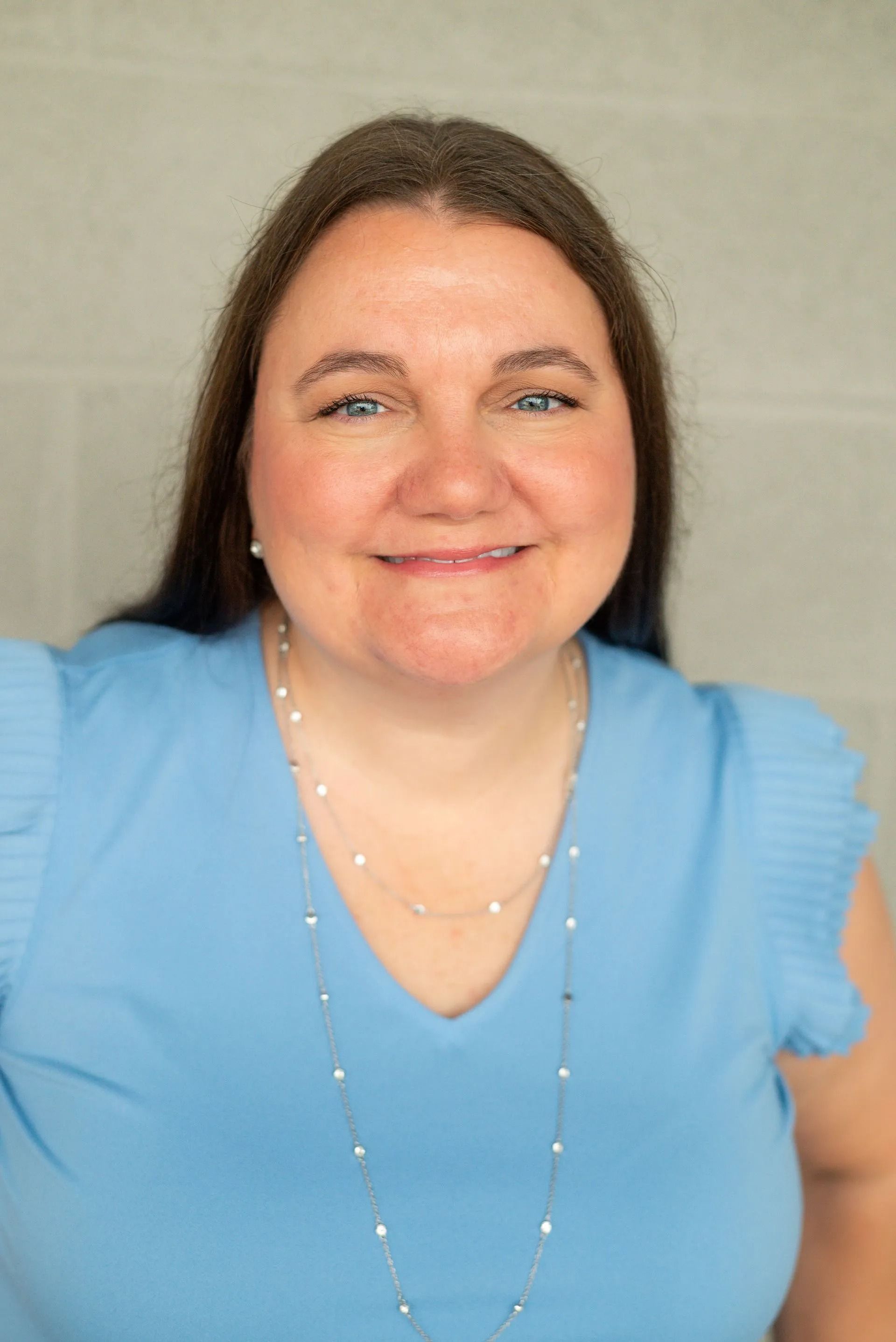 A smiling woman with long brown hair wearing a light blue, short-sleeved blouse and a thin, silver necklace.