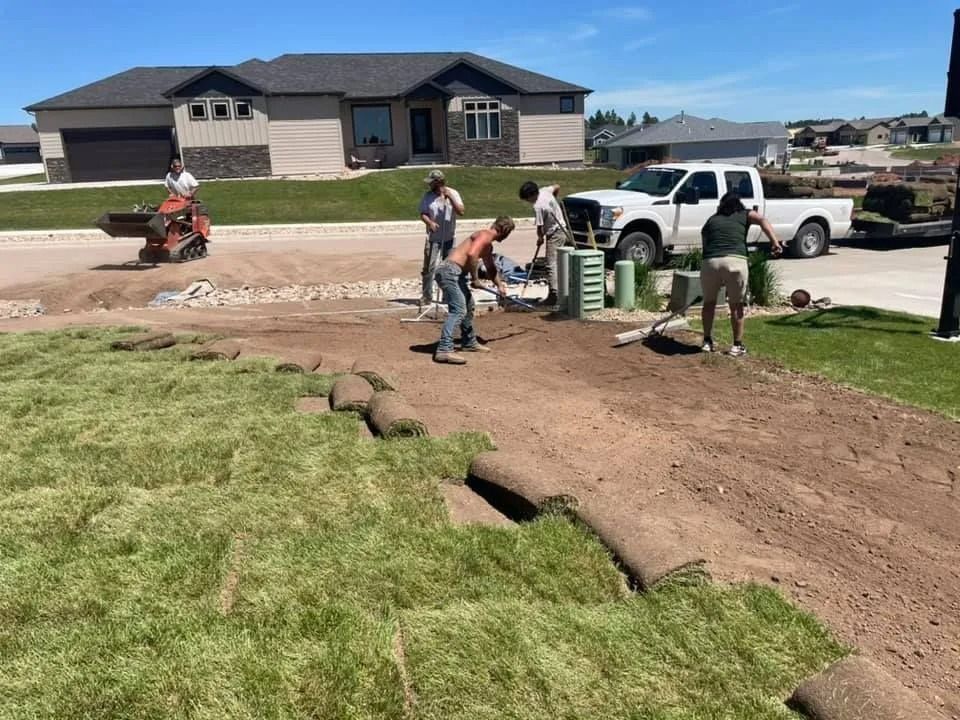 A crew lays sod on a dirt lawn in a residential neighborhood with a truck and equipment nearby on a sunny day.
