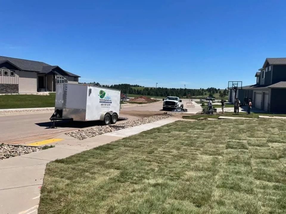 Landscapers install new sod on a sunny day in a suburban neighborhood with a white trailer parked nearby.
