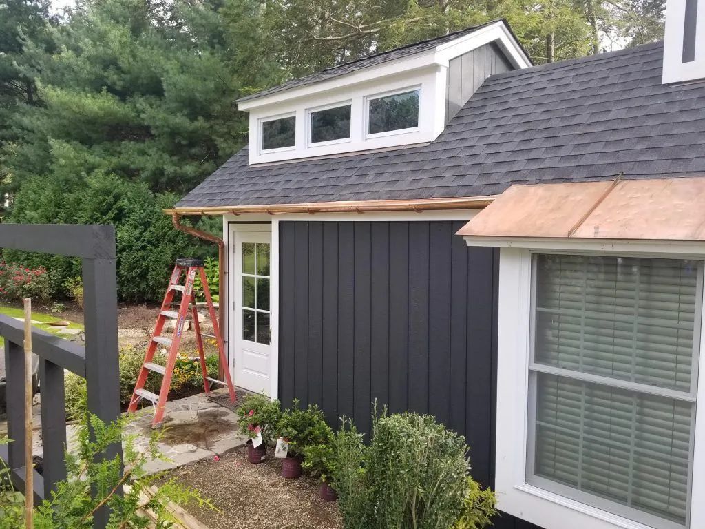 Dark gray building with copper gutters, a white door, and a three-window dormer. A red ladder leans against the wall.