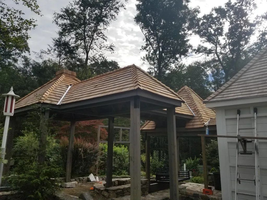 Wooden gazebo with brown roof, surrounded by trees. A white building with a ladder is on the right.