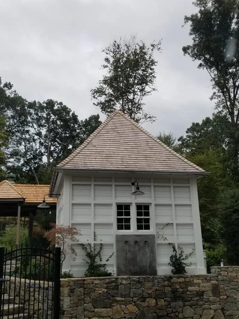 White building with a gray roof and grid-like facade on a stone wall, surrounded by trees.