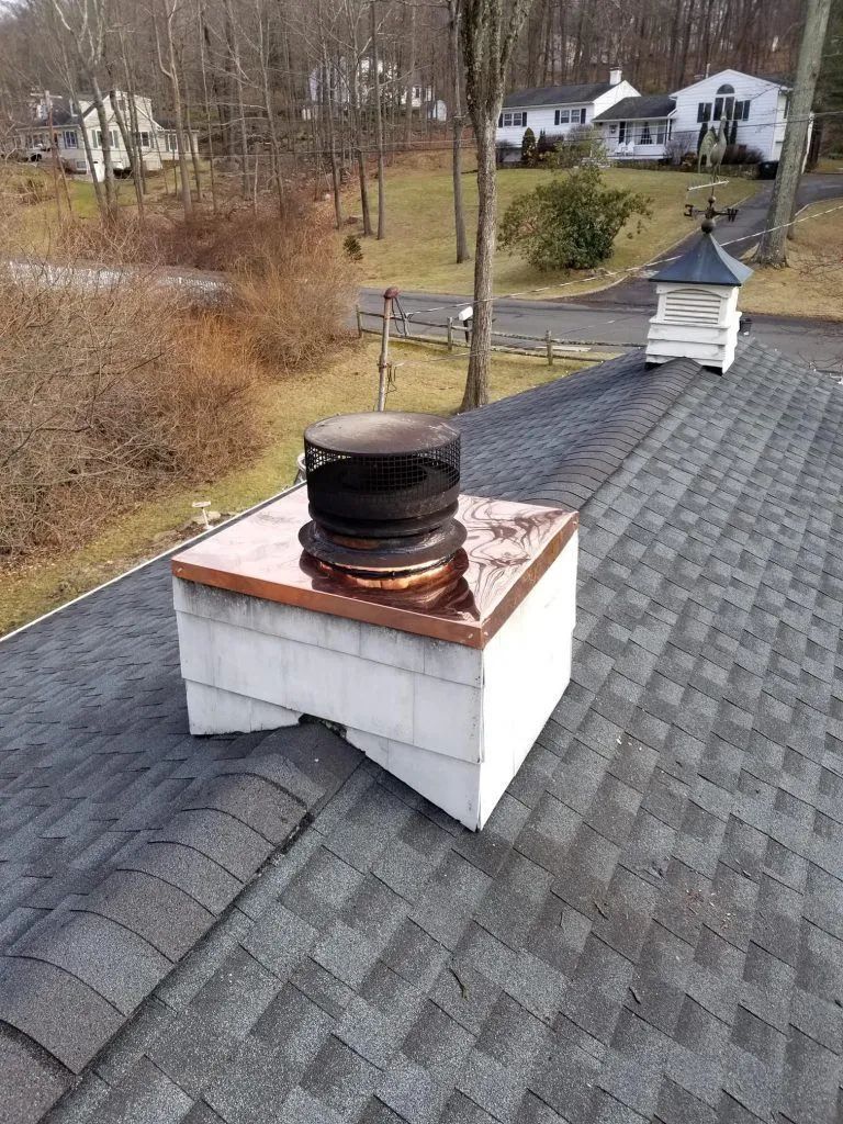 Chimney on a gray shingle roof with copper flashing, topped with a black metal flue cap.