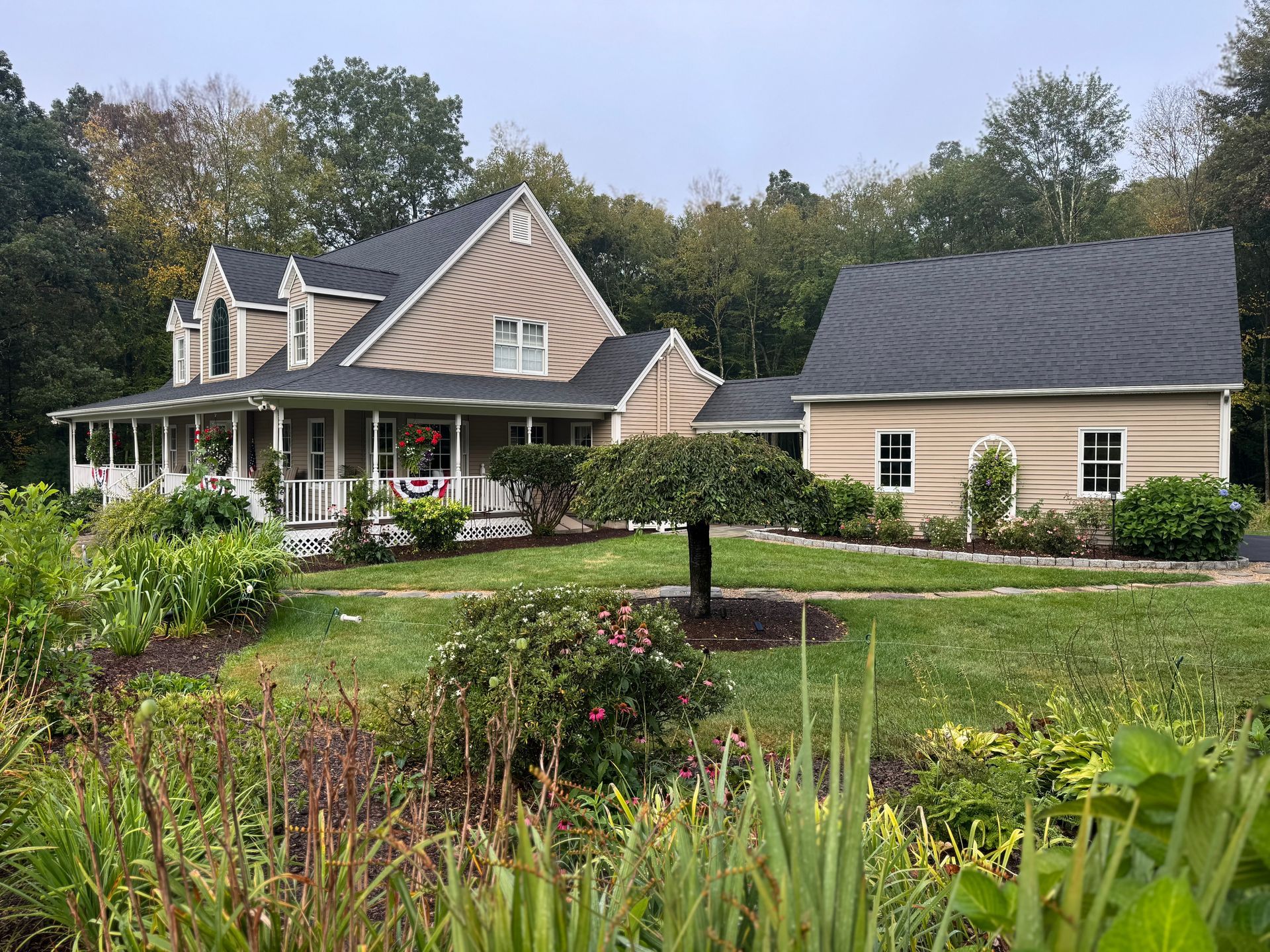 Tan house with porch and a detached building on a green lawn with a garden in the foreground.
