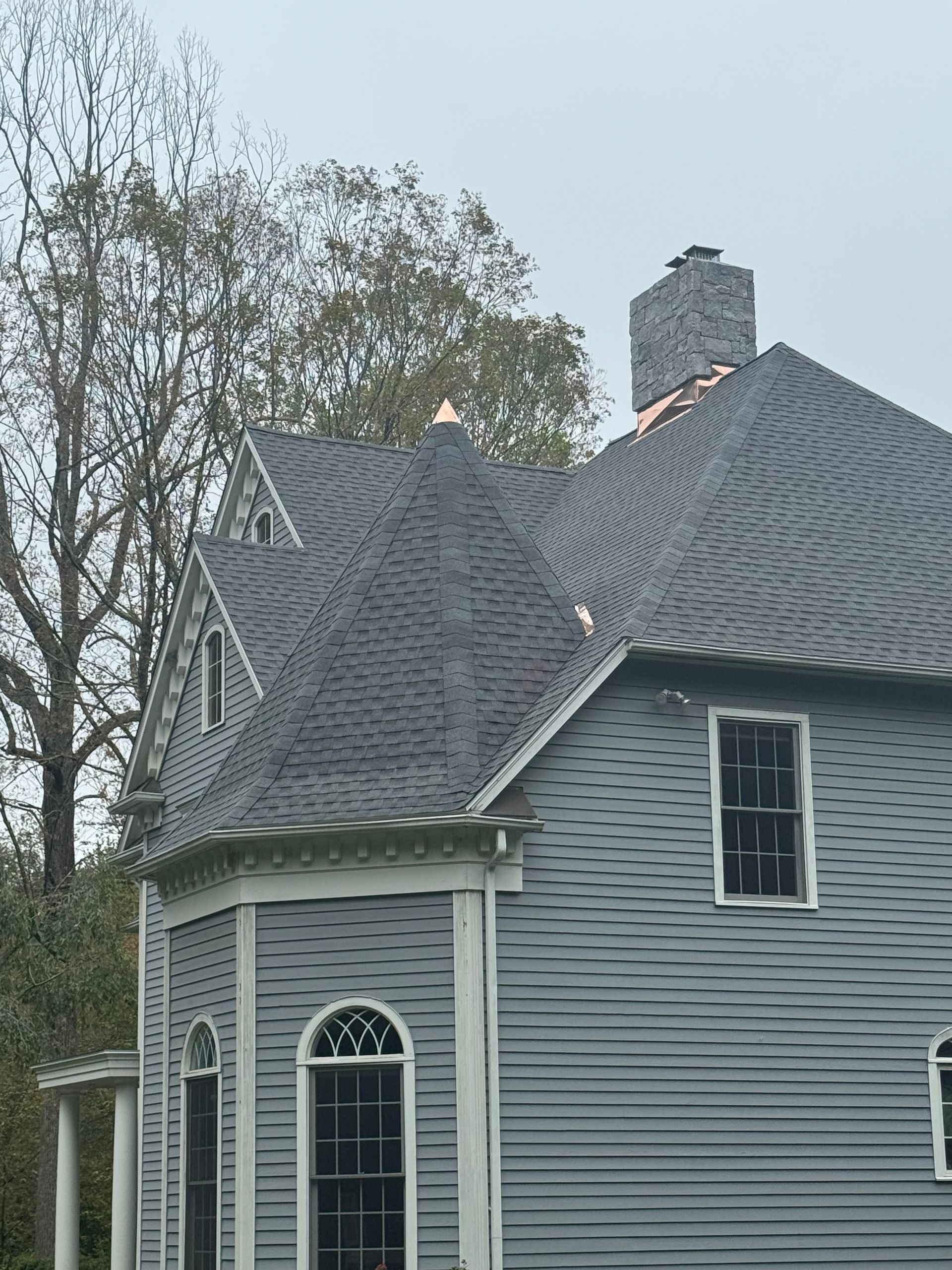 Gray house with dark gray shingle roof, chimney, windows, and trees in the background.