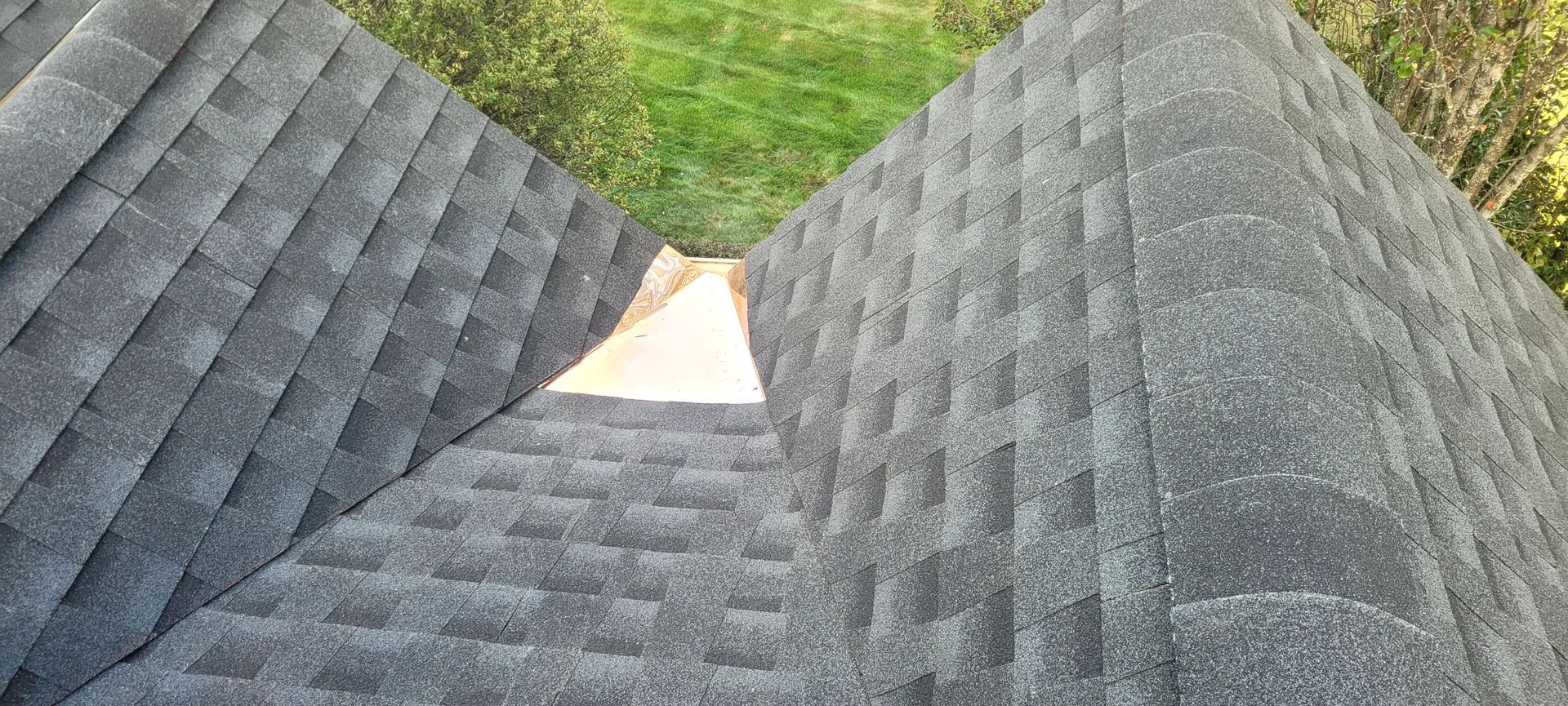 Overhead view of a roof with gray asphalt shingles and a valley, green foliage in background.