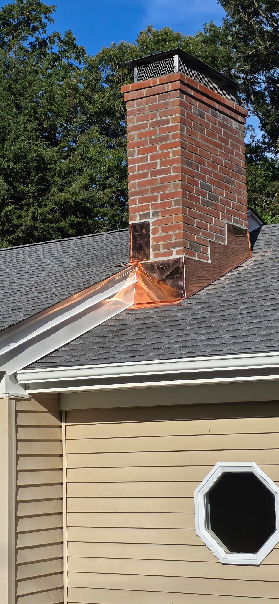Brick chimney with copper flashing on a roof, surrounded by trees and a blue sky.