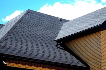 Dark asphalt shingle roof on a house, with black gutters and tan brick wall.