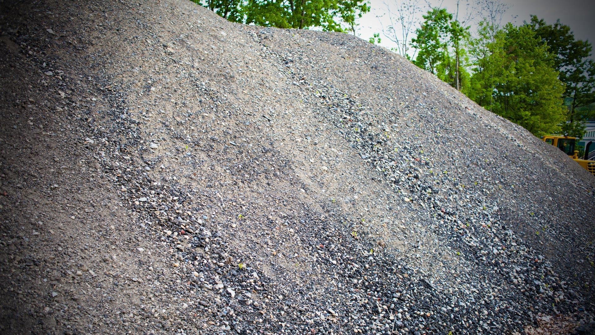 Large pile of gray gravel or crushed stone outdoors, with trees visible in the background.