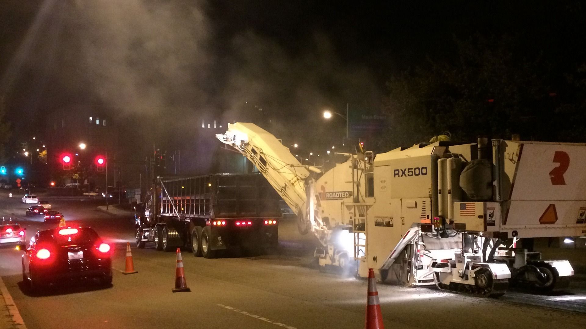 Night road construction with workers, traffic cones, flashing lights, and a large paving machine emitting steam.