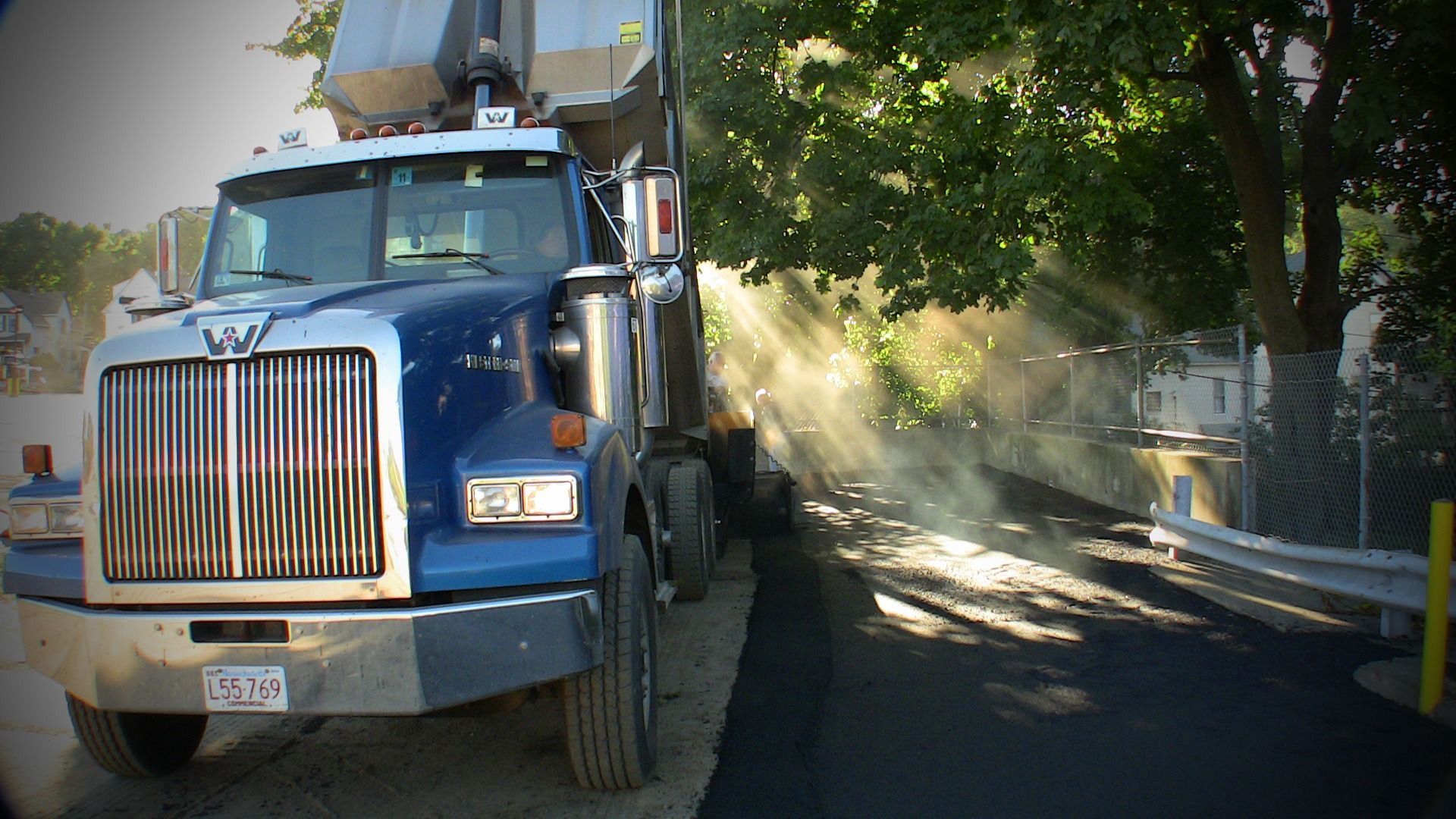 Blue dump truck driving on a tree-lined street with dust or debris in sunlight