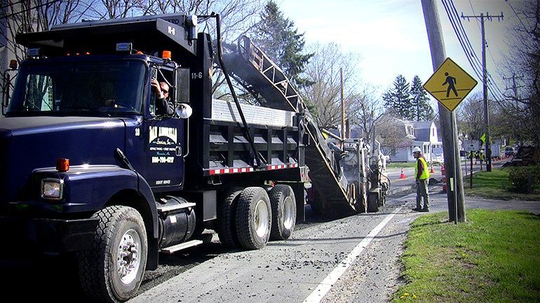 Roadwork truck paving a street while a worker stands beside a sidewalk on a sunny day