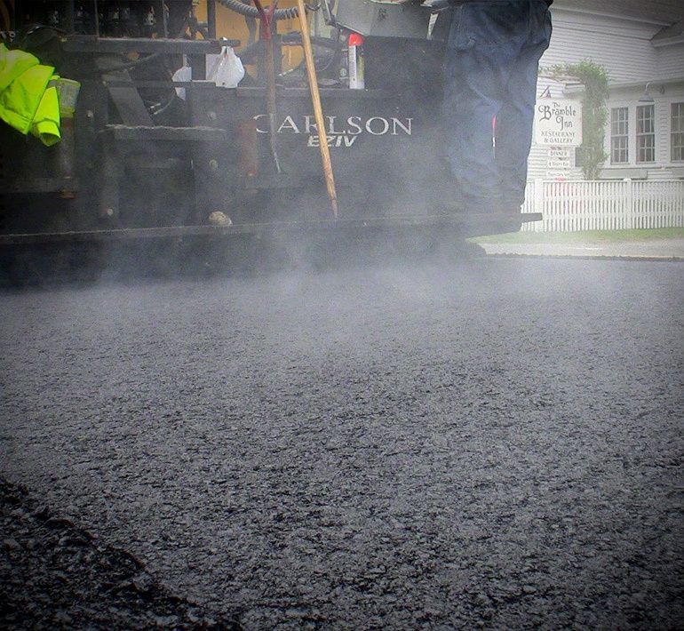 Black asphalt being laid with steam rising, near a house and a truck.