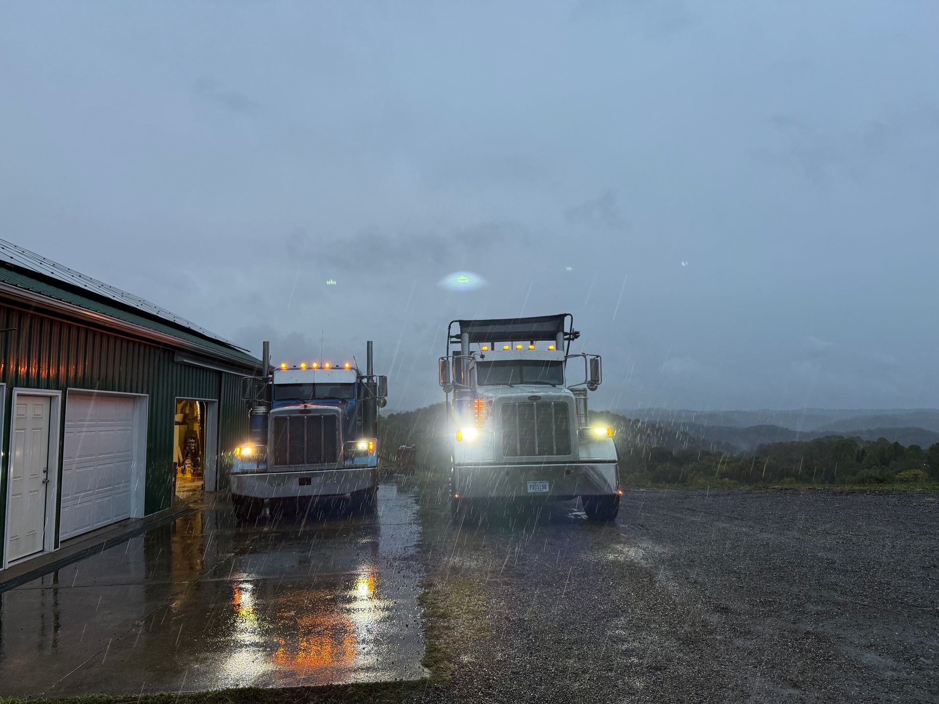 Two semi-trucks parked near a building in the rain, headlights on, with a dark sky.