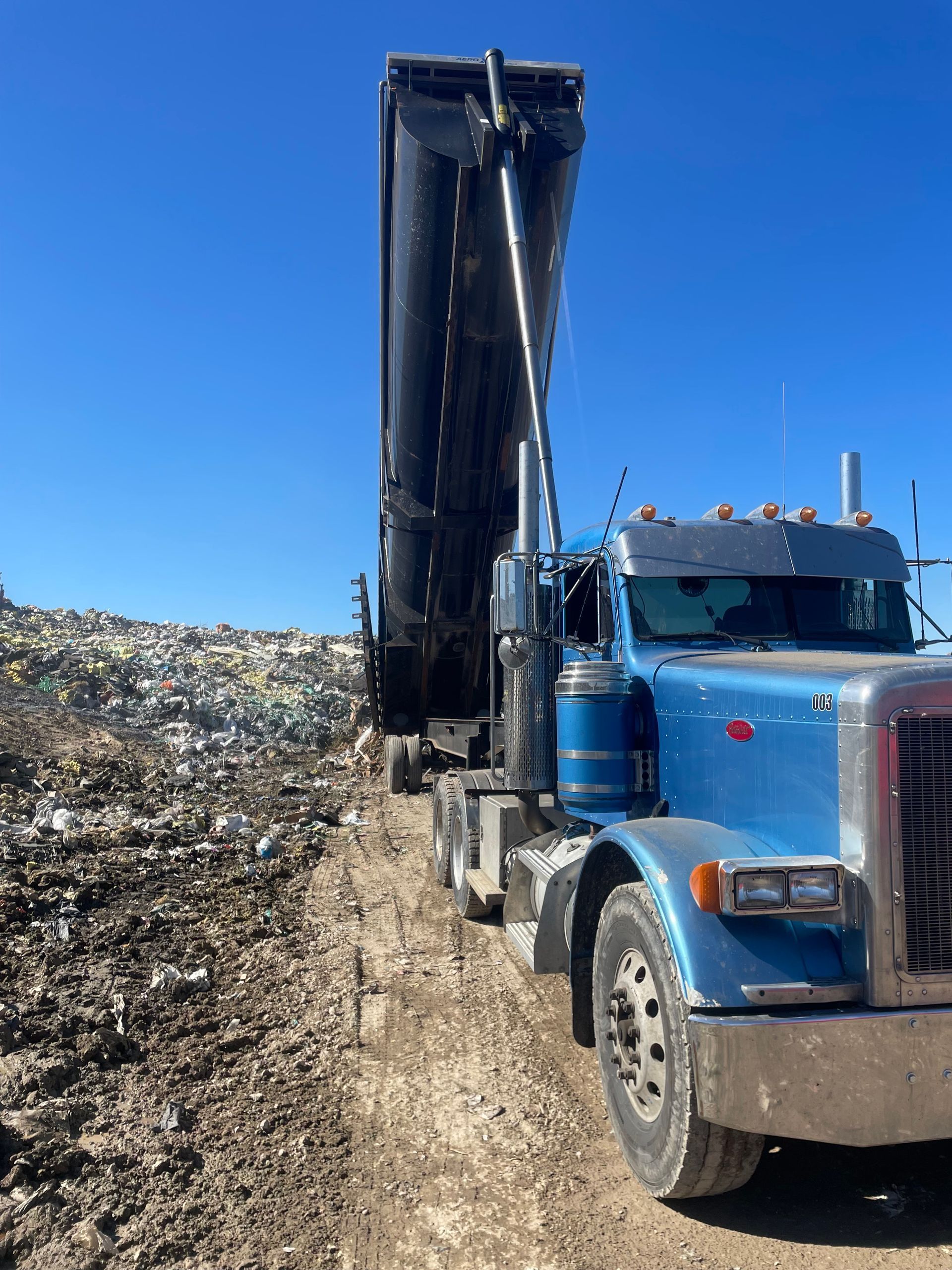 Blue dump truck unloading at a landfill under a clear blue sky.