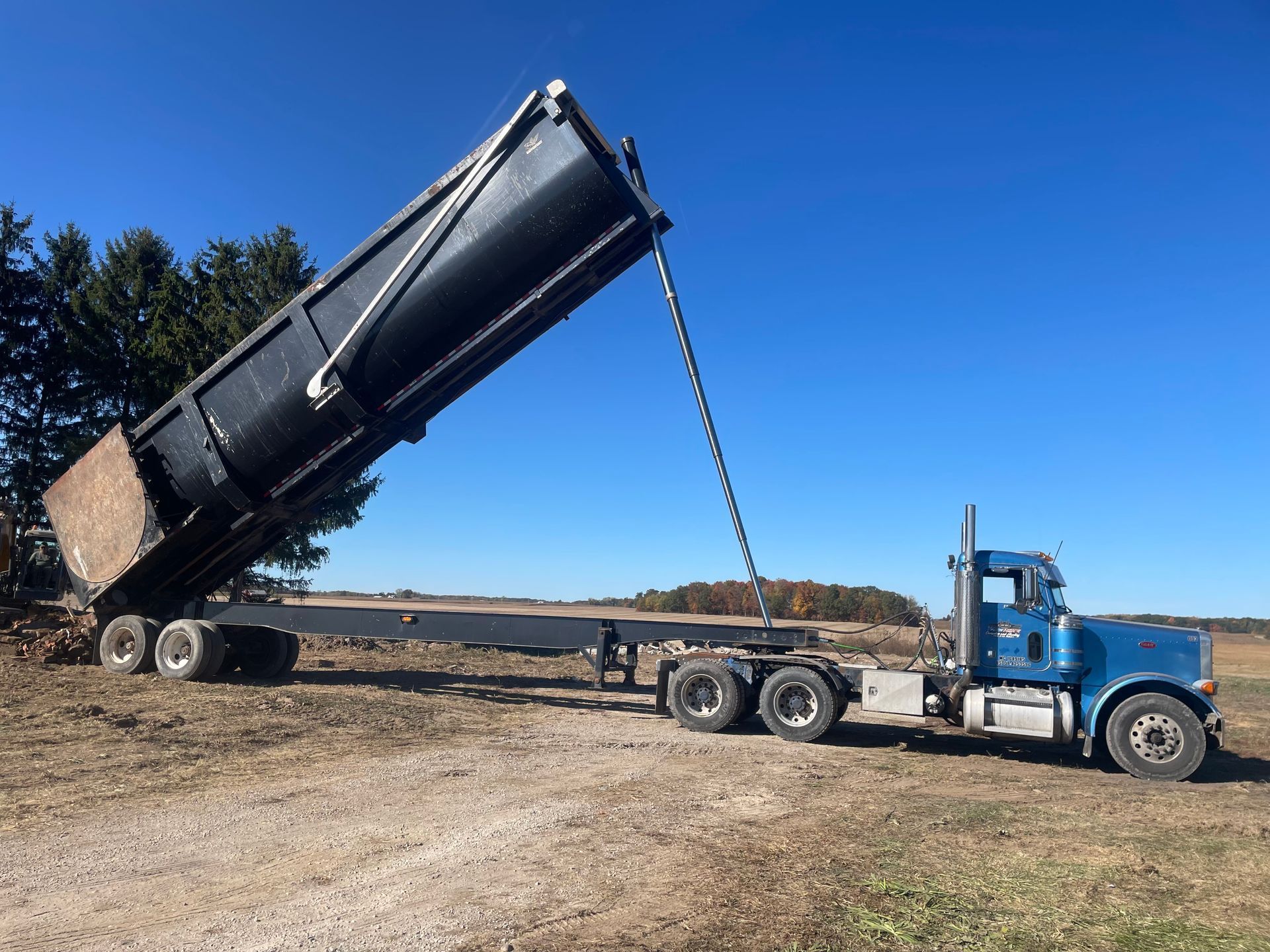 Blue semi-truck with a black dump trailer raised in a field under a clear, blue sky.