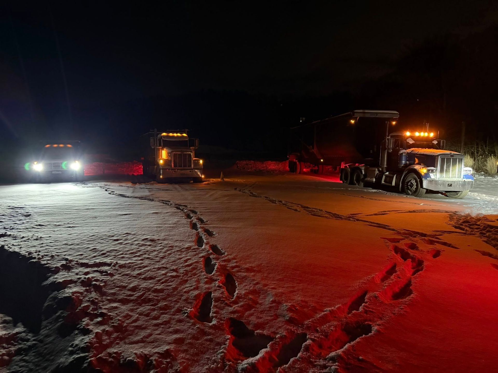 Three trucks illuminated at night on a snowy area. Footprints lead towards the trucks.