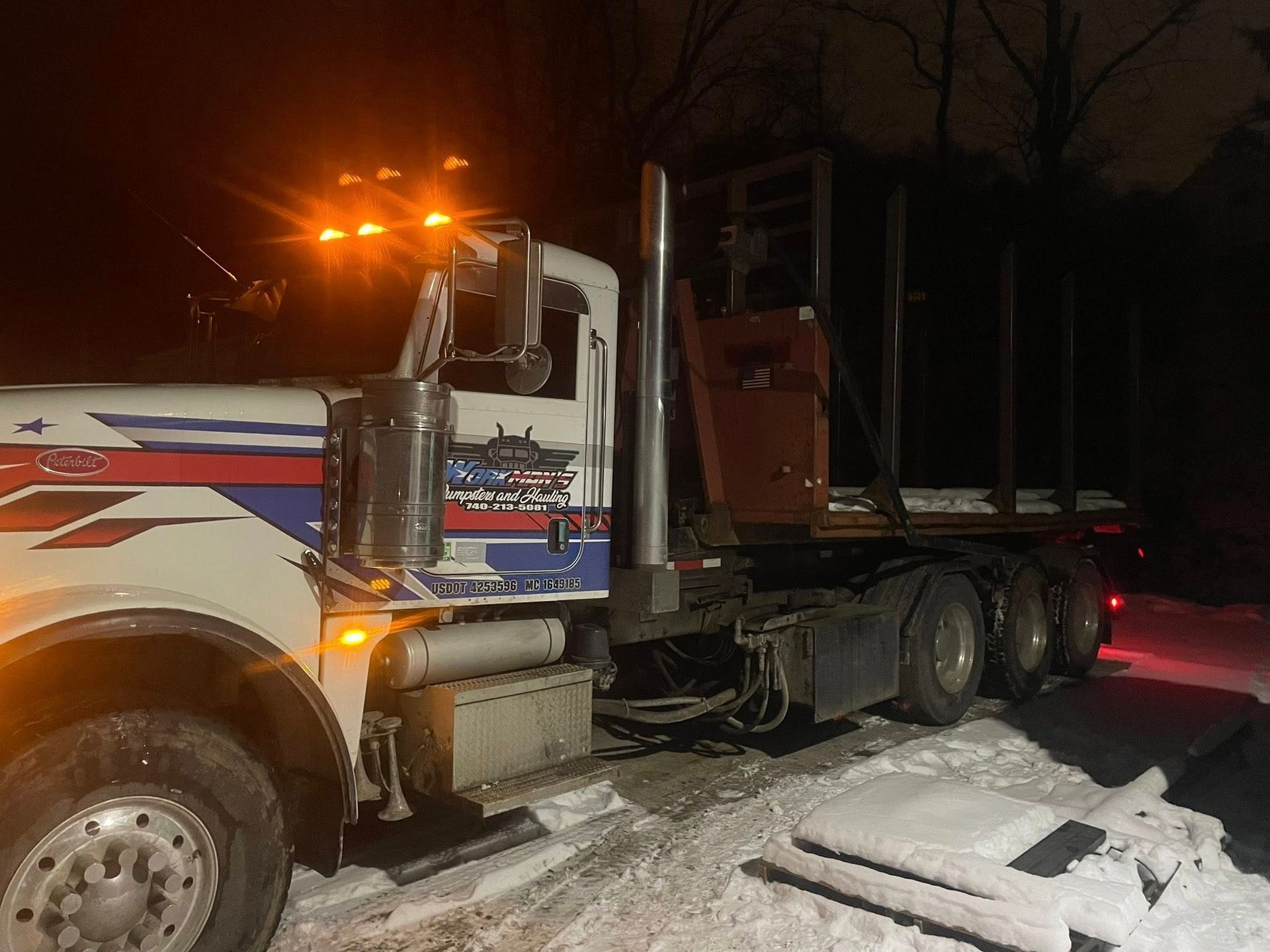 White semi-truck with a log trailer parked on a snow-covered road at night; lights are on.