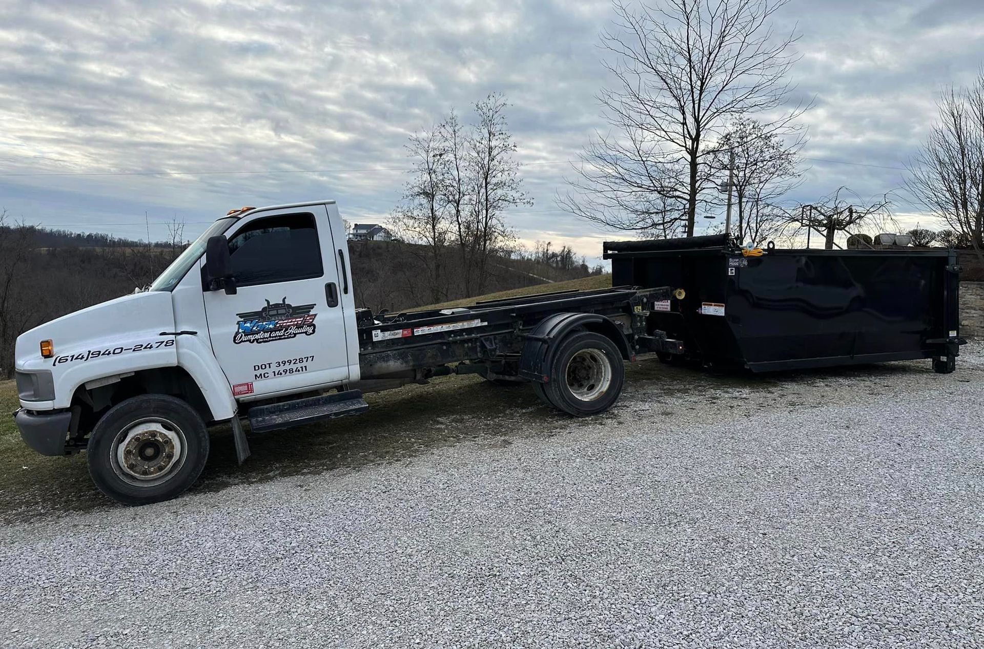A white dump truck is parked next to a black dumpster in a gravel lot.