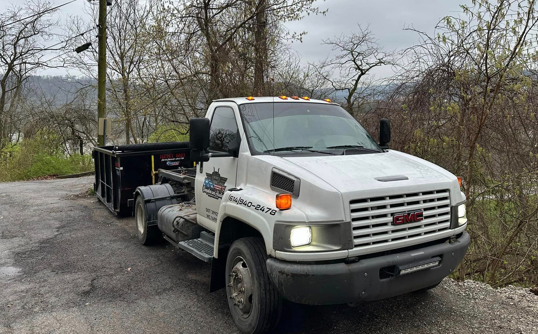 A white tow truck with a dumpster attached to it is parked on a gravel road.
