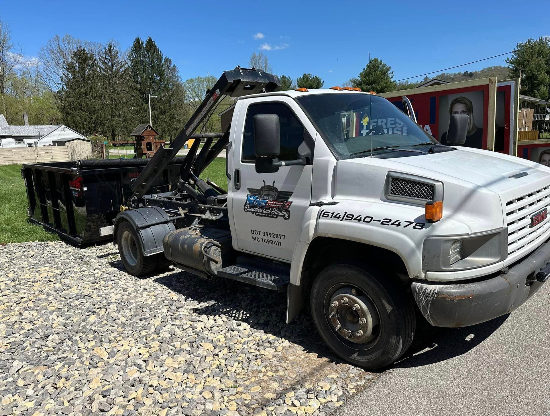 A white tow truck with a dumpster attached to it is parked on a gravel road.