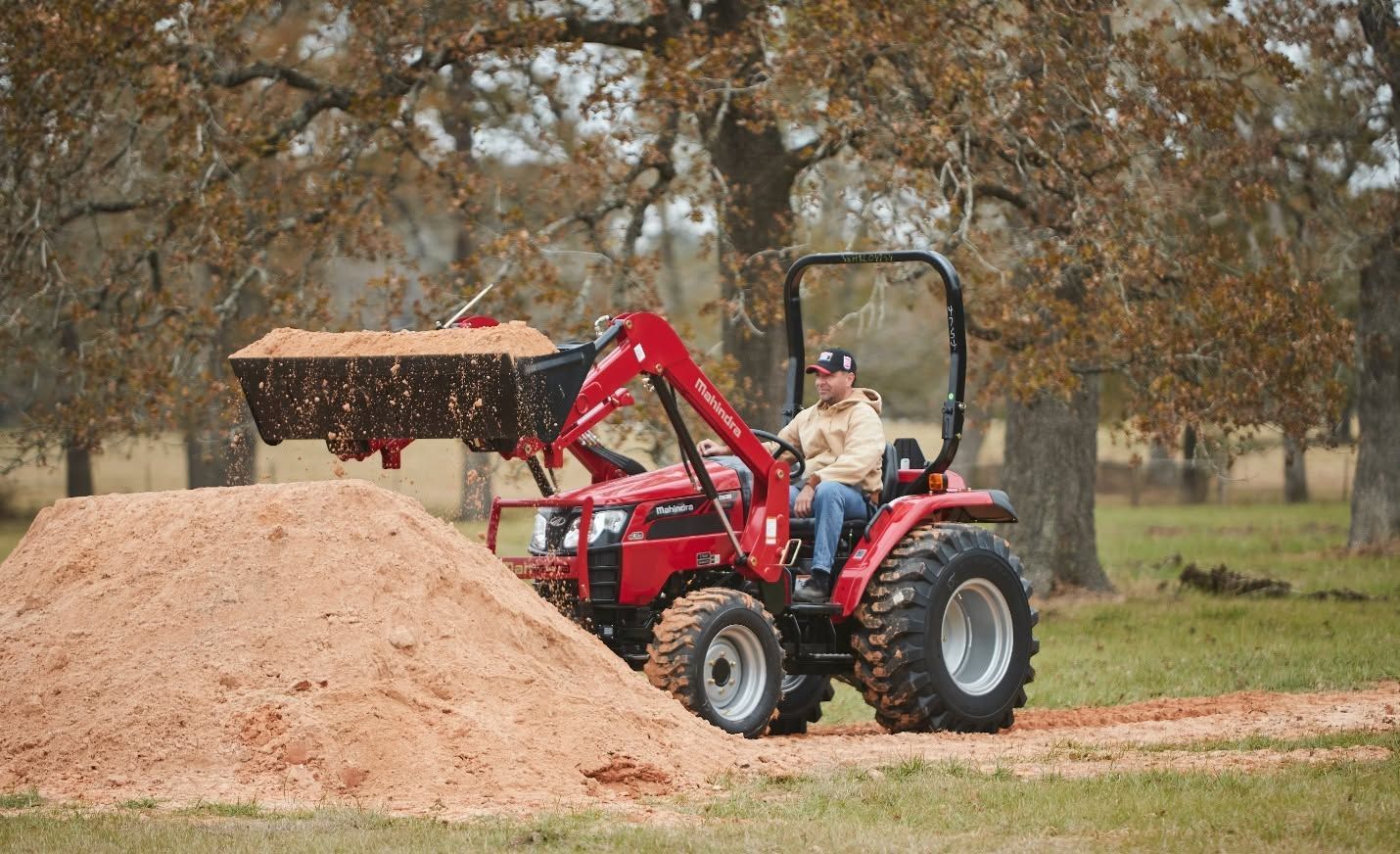 Red tractor with front loader dumping mulch beside a large pile in a wooded field