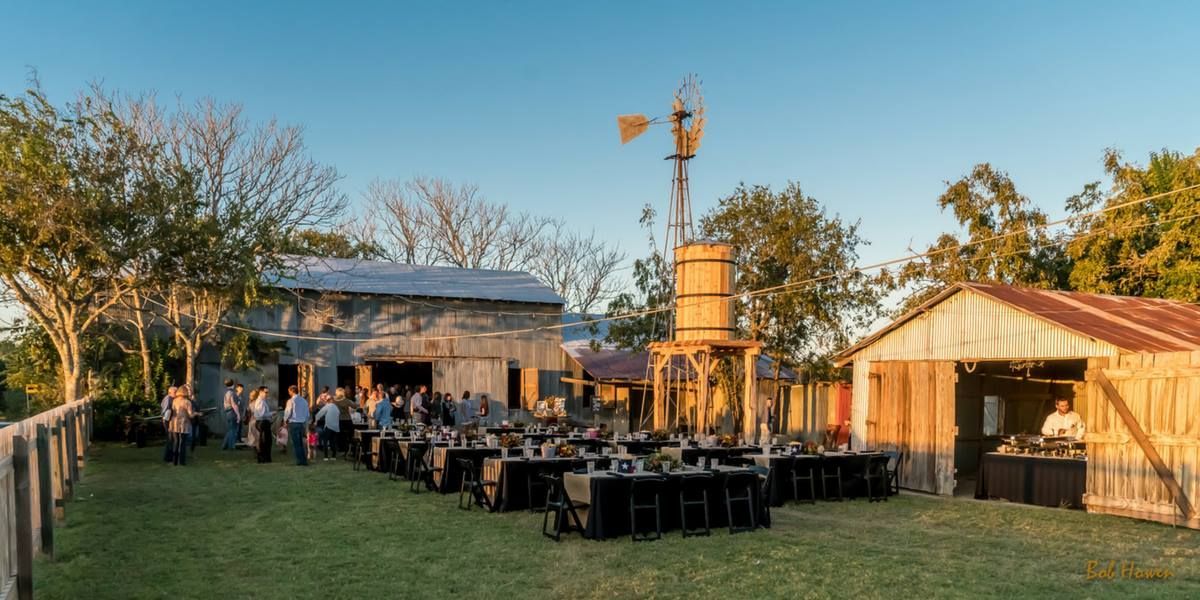 A group of people are sitting at tables in a field in front of a barn.