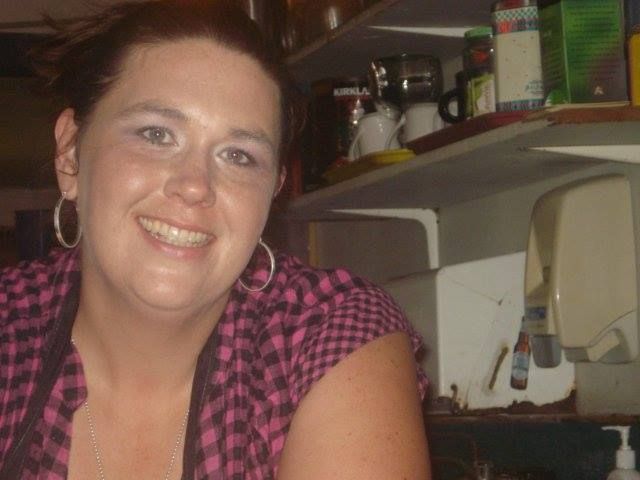 A woman in a pink plaid shirt is smiling in the kitchen.