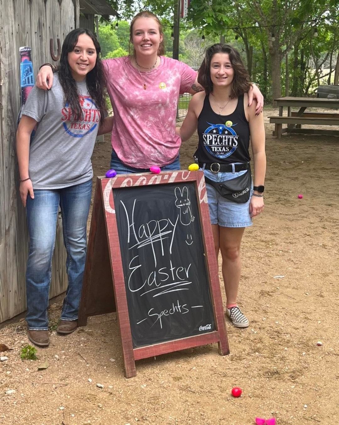 Three women are posing for a picture in front of a sign that says happy easter spirits.