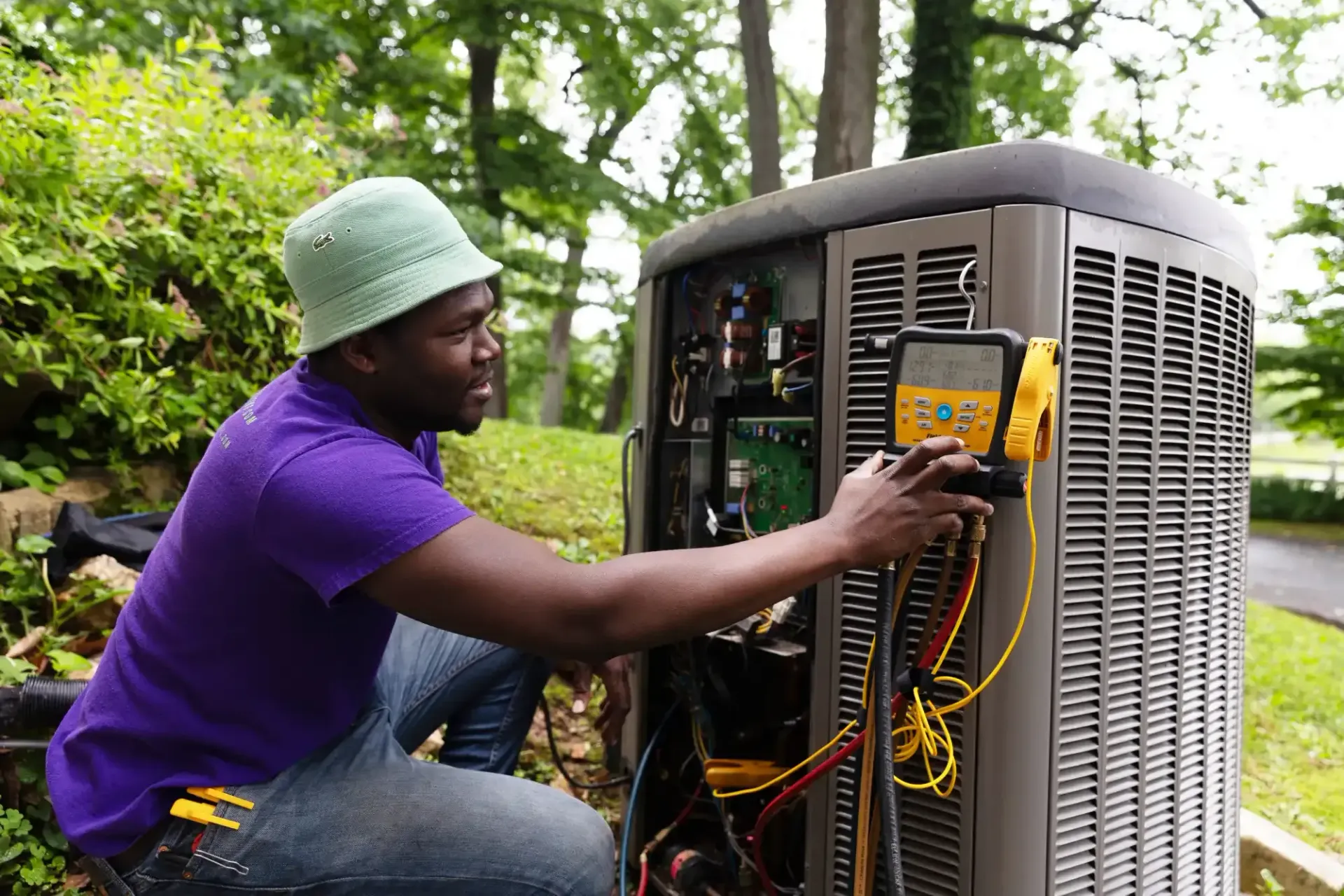 Technician in purple shirt and green hat repairs an outdoor air-conditioning unit.