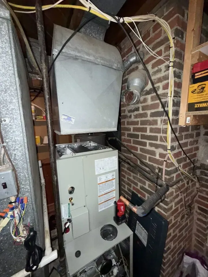 Open furnace and HVAC unit in a cramped brick basement with exposed wires and ducts